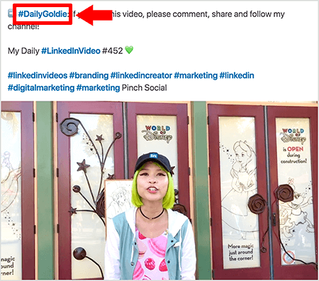 هذه لقطة شاشة توضح كيف تستخدم Goldie Chan علامات التجزئة في نص مشاركات الفيديو الخاصة بها على LinkedIn. تشير وسائل الشرح الحمراء إلى علامة التصنيف #DailyGoldie في النص ، والتي تعتبر فريدة في منشورات الفيديو وتساعدها في تتبع المشاركات. يتضمن المنشور أيضًا علامات تصنيف أخرى ذات صلة تساعد الأشخاص في العثور على الفيديو الخاص بها ، بما في ذلك #LinkedInVideo. في صورة الفيديو ، تقف جولدي أمام بعض الأبواب في إحدى شاشات عالم ديزني. إنها امرأة آسيوية ذات شعر أخضر. إنها ترتدي قبعة لينكد إن سوداء ، وقلادة عنق سوداء ، وقميص وردي بطبعة ماكرون ، وسترة زرقاء وبيضاء.